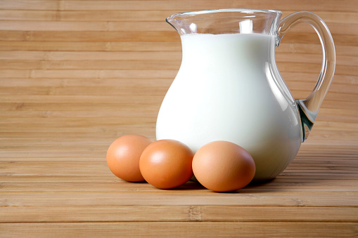 A DSLR photo of a glass pitcher of milk together with three eggs on a wooden bamboo background. The side view, space for copy.