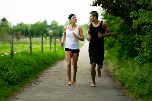 Young couple jogging outdoors in bare feet. They are running down a path lined with trees on one side and a fence on they other. Horizontal shot. Note to inspector focus on foreground some motion blur.