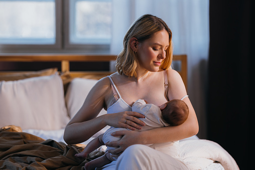 A smiling Caucasian mother feeding her cute baby while holding it. She is sitting on the bed in her bedroom.