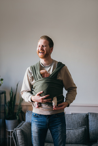 A smiling Caucasian father carrying his cute baby in a ring sling. He is standing in the living room.