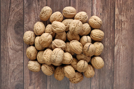 Stock photo showing close-up, elevated view of a pile of unshelled walnuts against a brown, wood grain background. Raw walnuts are considered to be a very healthy snack food, containing a wealth of essential Omega-3 fatty acids, antioxidants and protein, and boasting a list of health benefits while reducing stress.