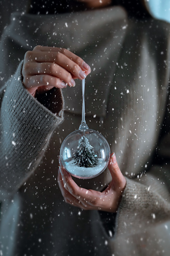 Woman in grey sweater holding Christmas tree toy glass decorative ball in hands while snowing, copy space, Christmas, new year holiday celebration concept