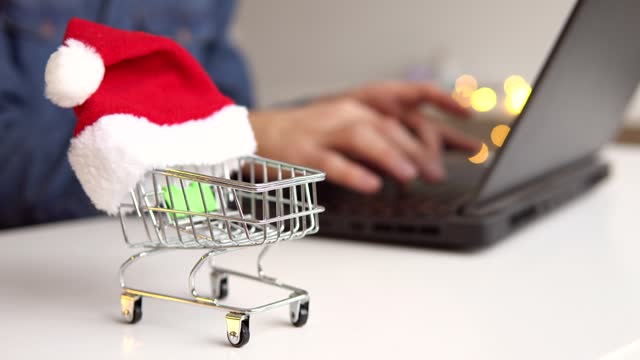 Shopping cart with small santa hat on table. Man using laptop on the background. Shopping online during holidays.