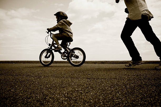 Pai perseguindo jovem ciclista aprender a surfar - foto de acervo