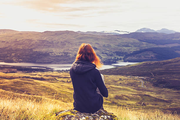 Rear view of a young woman sitting on a rock on top of a mountain and admiring the sunset