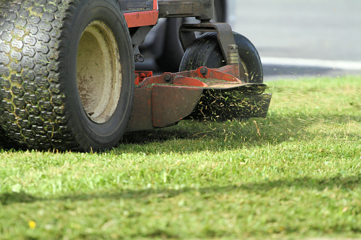 Close-up shot of a riding mowers discharge chute with focus on grass cuttings being ejected. Very shallow dof and motion blur on tires.