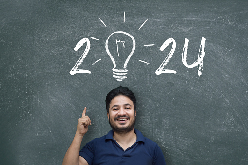 Indian/Asian young man pointing up while standing against blackboard with chalk drawing of light bulb and new year 2024