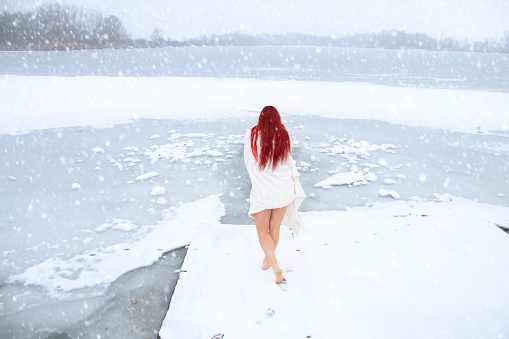 Rear view woman with long red hair and pretty legs walking outside barefoot on the snow to the icy lake for the natural ice bath while snowing. Hardening in cold water, winter open water swimming and healthy cryotherapy concept