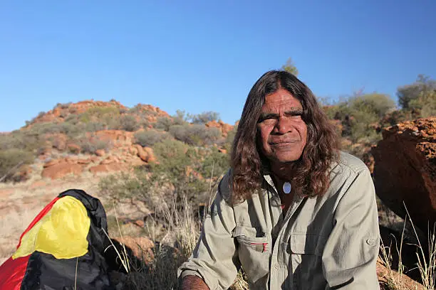 An Indigenous Elder sitting in the outback with the Aboriginal flag in the back ground