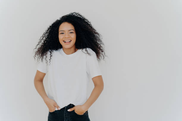 Laughing Black woman with curly hair in a white shirt and jeans against a plain background