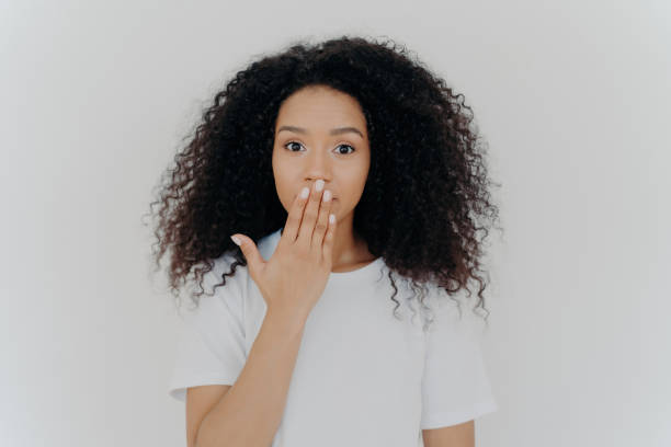 Surprised Black woman with curly hair, hand over mouth, white tee, plain backdrop