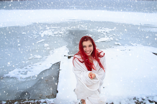 Happy young woman with long red hair, wrapped only in a blanket with a joyful smile enjoys natural cold therapy in winter outdoors and healthy lifestyle, hardening on snow, ice bath at the natural lake in snowy wild nature