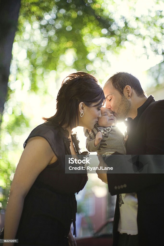 Young Hispanic Family In Urban Park A young adult family kiss their baby boy in a metro city park, the sun shining through the green trees behind them.  Vertical with copy space; very shallow depth of field. 0-11 Months Stock Photo