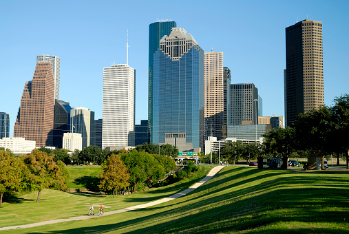 Horizontal, color, Skyline image of Houston, Texas seen across city park with two cyclists (a male and female) riding on a concrete path away from the city.  Backdrop of a beautiful clear blue sky.  Long shadows from trees are present in the lower right foreground in this late afternoon shot.