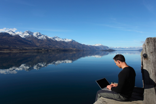 Man sitting by the lake typing on his computer. Beautiful outdoor location with mountain reflections. Plenty of copy space.