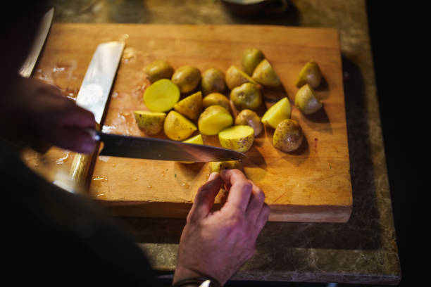 Detailed shot of a person's hand thinly slicing a fresh potato with a sharp steel knife on a dark wooden surface. The slices are stacked, emphasizing the prep work for a homemade meal. Suitable for concepts of healthy eating, cooking instruction, or potato recipes.