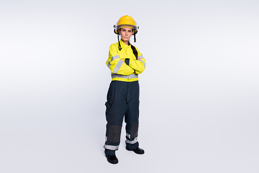 Beautiful woman working in construction in front of black background wearing blue work vest, white helmet and safety glasses. She is standing and posing for the camera with a smile. The girl was shot alone. The girl is wearing a white shirt and an industrial uniform. She stands smiling.
