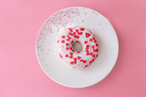 White donut sprinkled with small pink hearts placed on a beautiful white plate with pink flowers. Pink background.