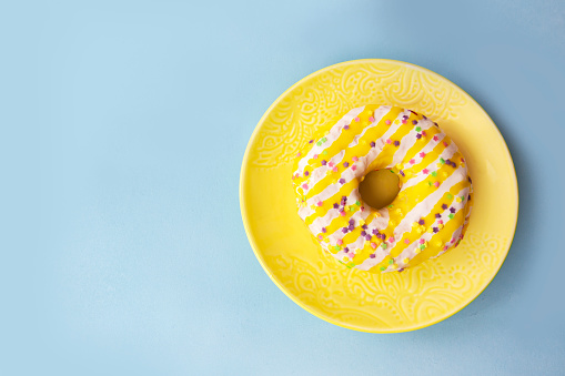 Donut with white - yellow icing and sprinkled with small colored flowers laid on a yellow plate. Blue background.
