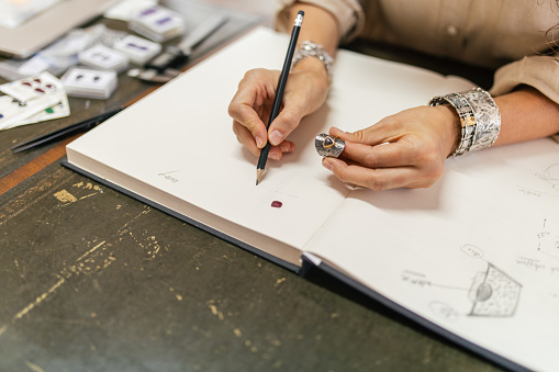 Mature adult woman sitting in her manufacture workshop and designing jewelry. She is drawing in her sketchbook. She is a designer and artist of unique jewelry made of precious metal. She is doing all process phases from design, manufacture and quality control.