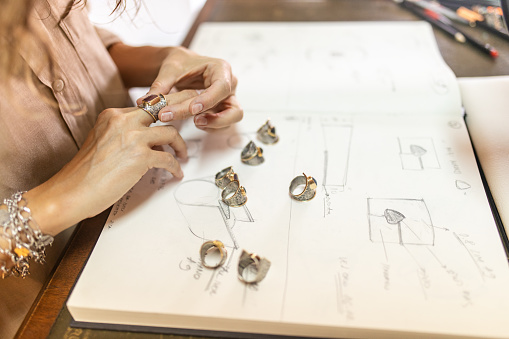 Mature adult woman sitting in her manufacture workshop with her sketchbook of jewelry open in front of her. She is putting one of the rings on her finger and looks like she is examining it. She is a designer and artist of unique jewelry made of precious metal. She is doing all process phases from design, manufacturing and quality control.