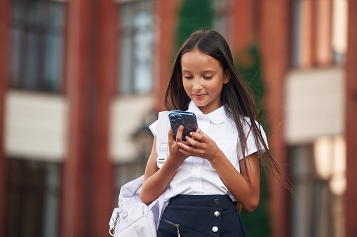 Using smartphone. School girl in uniform is outdoors near the building.