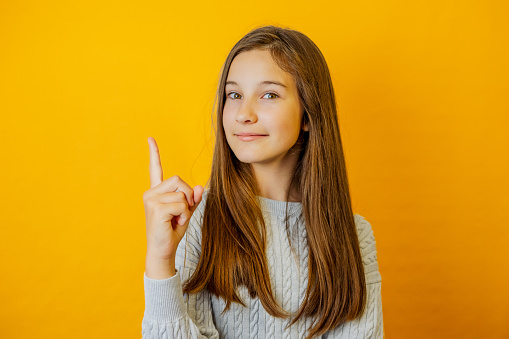 Cute little girl raising her index finger up in exclamation gesture and smiling at camera on isolated yellow background.