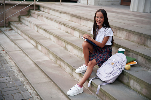Having free time, sitting on the stairs. School girl in uniform is outdoors near the building.