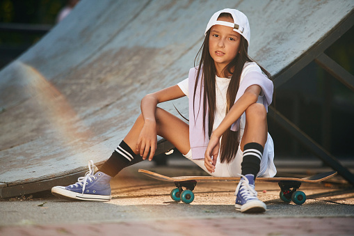 Taking a rest, sitting. Happy little girl with skateboard outdoors.