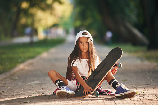 Front view, sitting on the ground. Happy little girl with skateboard outdoors.