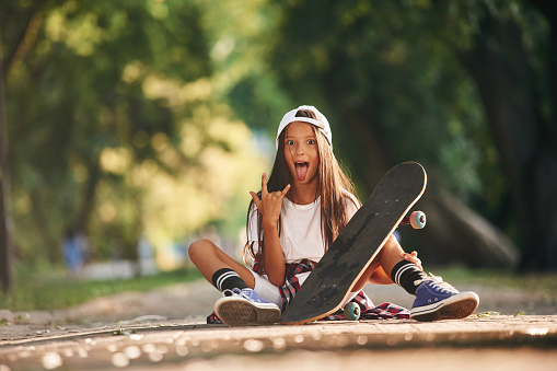 Showing metal cool hand gesture. Happy little girl with skateboard outdoors.
