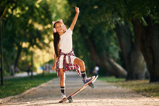 In cute pink headphones with ears. Happy little girl with skateboard outdoors.