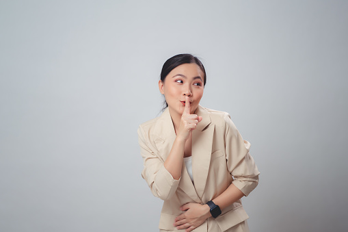 Asian woman happy smiling putting index finger on lips meaning keeping secret, standing isolated on white background.