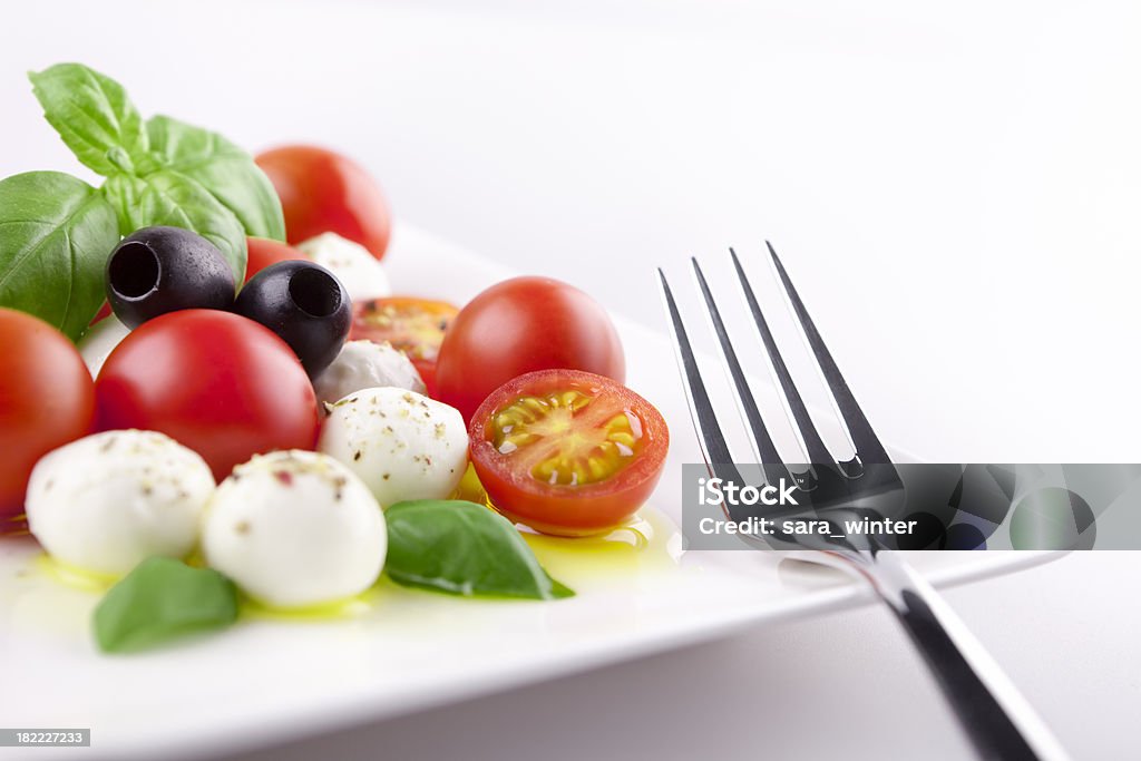 Fresh Caprese salad on a table in bright light "Caprese salad with mozzarella, cherry tomatoes, fresh basil, black olives and extra virgin olive oil. Shallow depth of field, focus on the cut tomato, mozzarella next to it, fork and olives." Caprese Salad Stock Photo