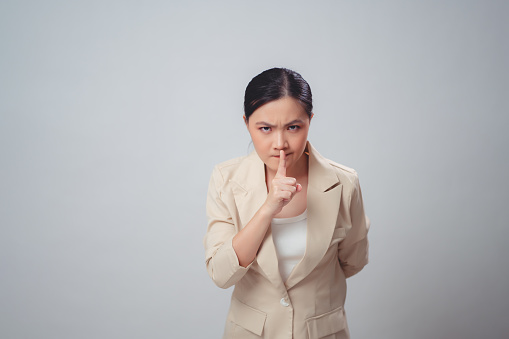 Asian woman annoyed frowning and putting index finger on lips meaning keeping silence, standing isolated on white background.