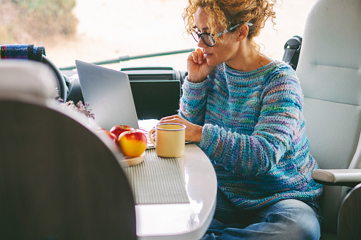 Adult nice woman work on laptop sitting in a camper van dinette enjoying freedom travel vacation or vanlife lifestyle. Modern job with computer connection technology. Female people freelance