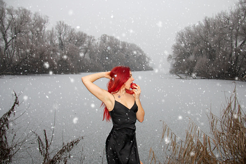 Fashion style woman in black dress eating the apple in a winter landscape with frozen lake while snowing, red hair contrasting with ice cold white nature