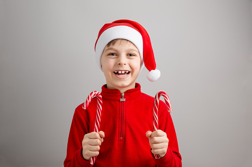 Cute boy in Santa Claus hat on isolated gray background showing striped lollipops, funny emotions. Merry Christmas concept.