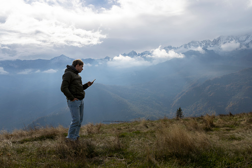 Young man uses mobile phone on mountain meadow above mountain range and lofty clouds