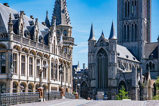 Stunning medieval Gothic architecture of Ghent, Belgium. The photo is taken from the St. Michael’s Bridge and shows the Belfry of Ghent and Saint Nicholas’ Church. The sky is a beautiful shade of blue and the buildings are illuminated by the sun. The intricate details of the buildings are visible, including the pointed arches, ribbed vaults, and flying buttresses.