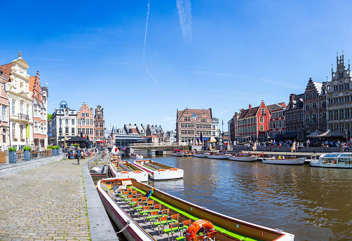 A canal in Ghent, Belgium. The canal is lined with colorful buildings on both sides, which are in the Flemish style with stepped gables and ornate facades. The boats on the canal are brightly colored and some of them are tour boats. Blue sky is on the background.