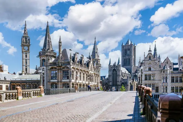 Stunning medieval Gothic architecture of Ghent, Belgium. The photo is taken from the St. Michael’s Bridge and shows the Belfry of Ghent and Saint Nicholas’ Church. The sky is a beautiful shade of blue and the buildings are illuminated by the sun. The intricate details of the buildings are visible, including the pointed arches, ribbed vaults, and flying buttresses.