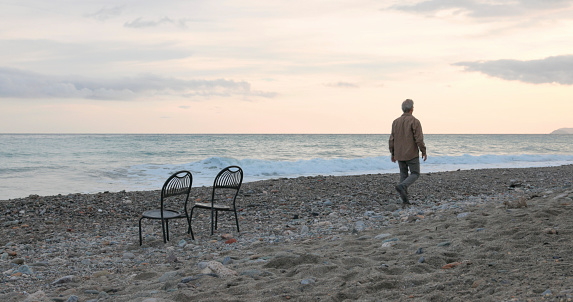 Senior man walks past chair on beach at sunrise and lofty clouds