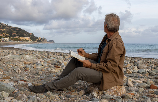 Senior man writes in journal on rocky beach