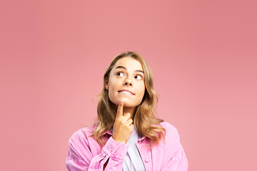 Portrait of attractive young girl choosing something looking up at copy space isolated on pink background. Shopping concept, discounts, sale