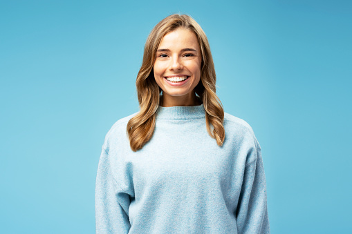 Smiling attractive woman wearing cozy sweater looking at camera standing isolated on blue background. Concept of beauty, makeup