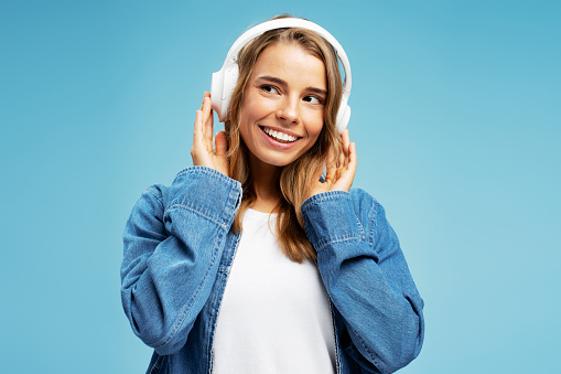 Portrait of attractive young woman wearing stylish casual clothes listening to music in wireless headphones looking away standing isolated on blue background. Technology concept, positive lifestyle