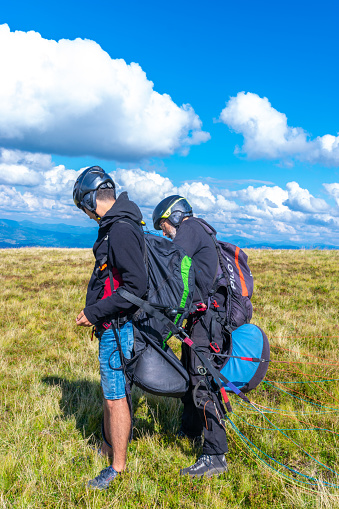 Pylypets, Ukraine - August 11, 2023: Young guy is preparing for a paragliding flight with an instructor. Beautiful summer sunny view from the top of the mountain. Blue cloudy sky.