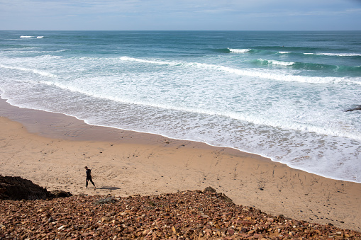 Aerial view of coastal headland, surfer walks along beach by waves, Lagos, Algarve