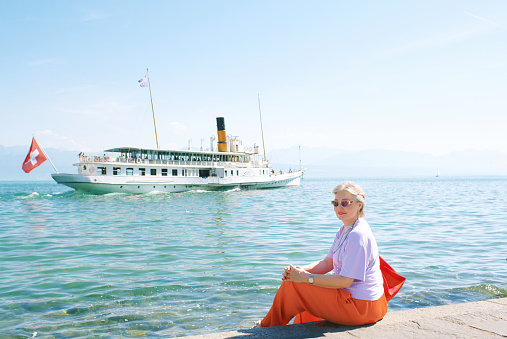 Outdoor portrait of mature 50 - 55 year old woman sitting by the lake, watching floating swiss french boat on lake Geneva, Switzerland